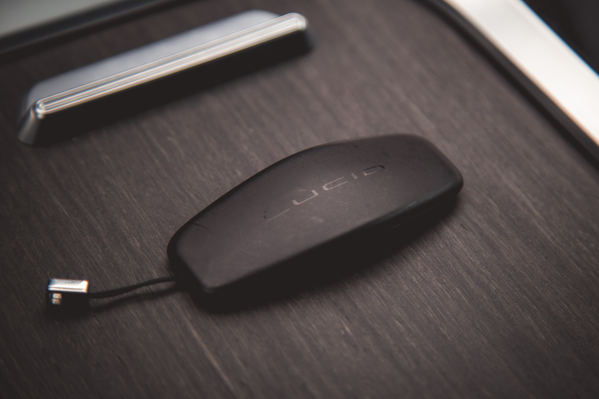 Una imagen en primer plano de un llavero negro con el logo 'Lucid', colocado sobre una superficie de madera oscura junto a un objeto metálico.