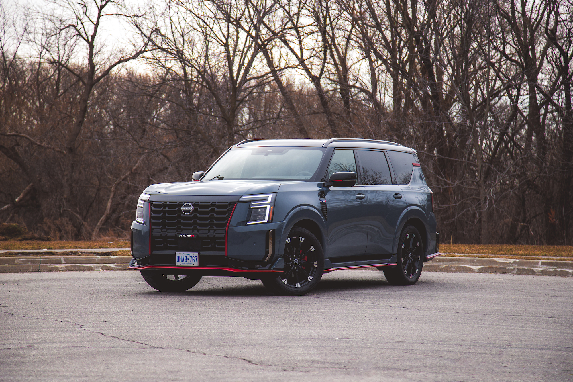 Un SUV Nissan gris estacionado sobre una superficie de asfalto con un fondo natural de árboles desnudos.