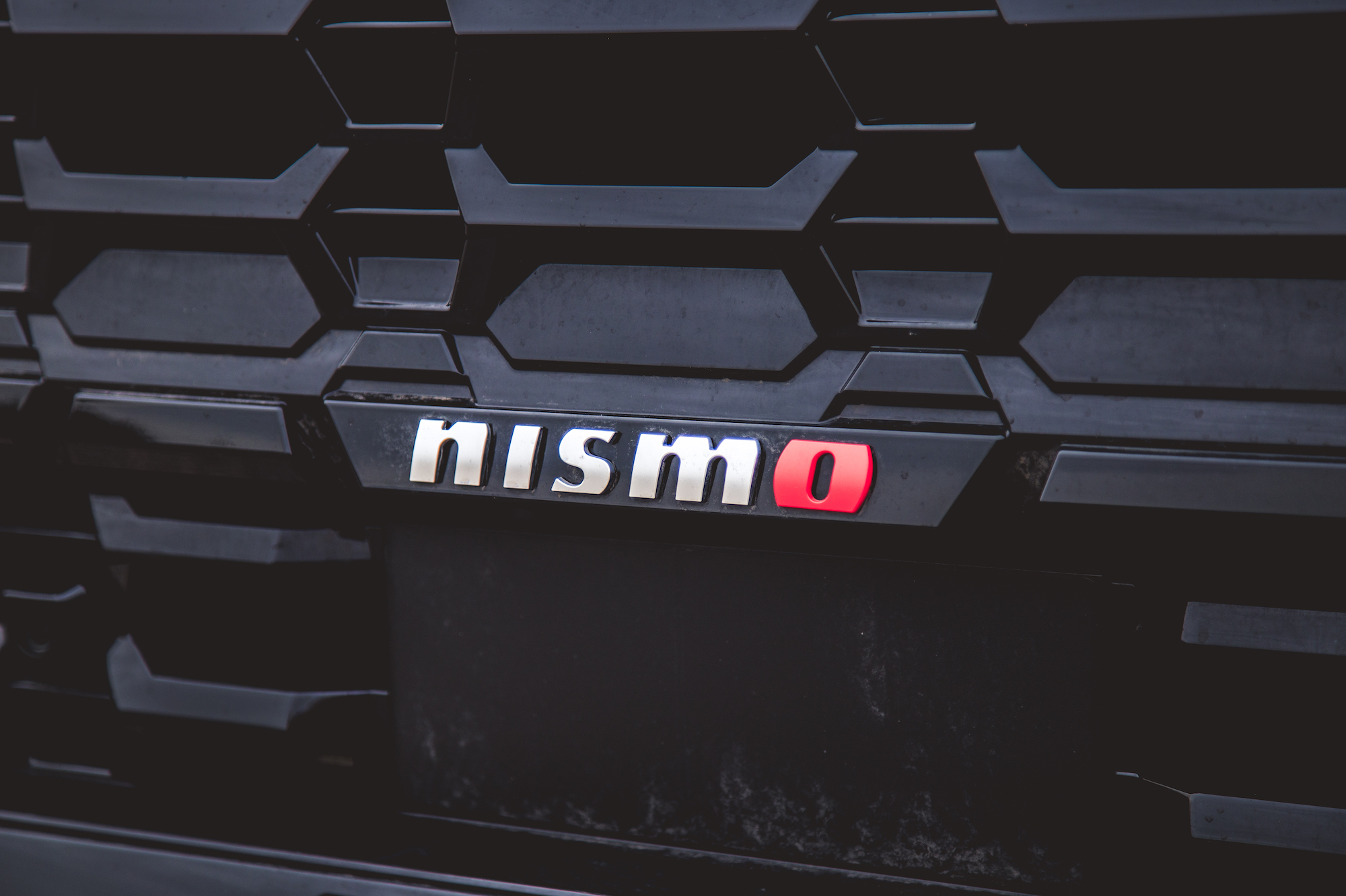 Primer plano de la parrilla de un vehículo negra con el logotipo 'Nismo' en plateado y rojo.