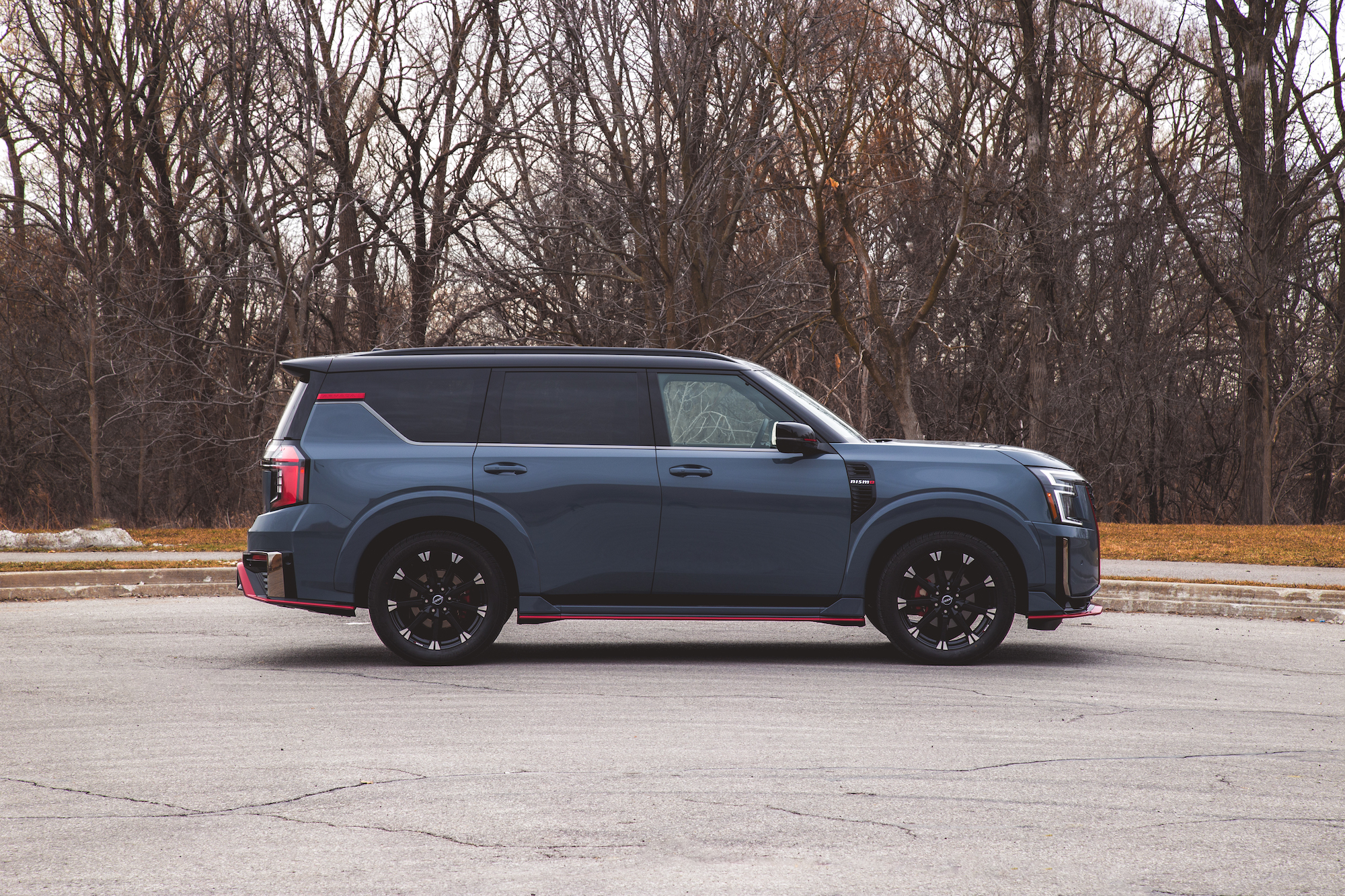Vista lateral de una SUV azul con un diseño elegante, ruedas negras y detalles en rojo, estacionada sobre una superficie de concreto con árboles desnudos al fondo.