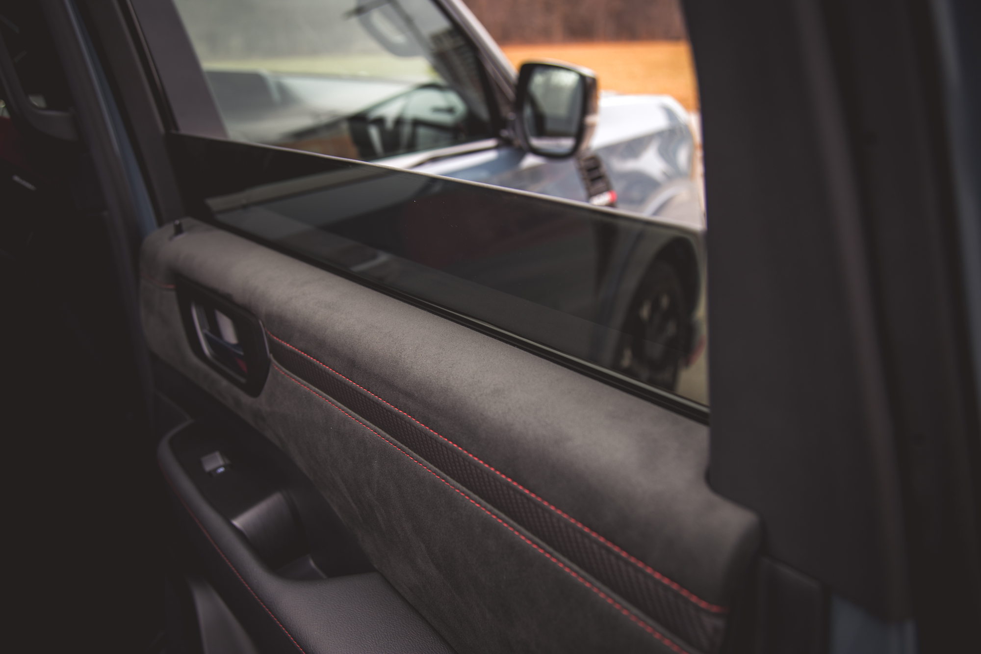 Vista interior de la puerta de un automóvil, con un reposabrazos de gamuza y cuero con costuras rojas y un elegante diseño de ventana.