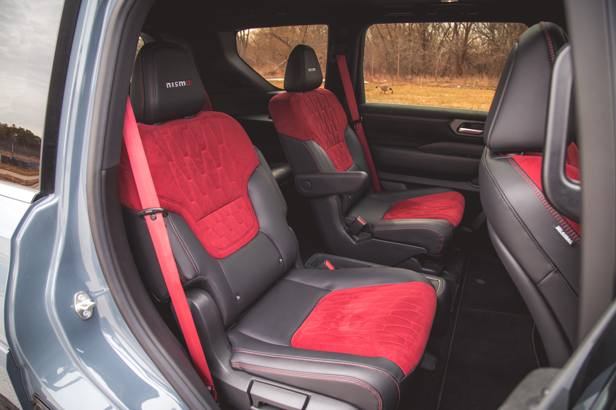 Vista interior de un automóvil que muestra dos asientos deportivos rojos y negros con la marca Nismo, con un fondo panorámico exterior visible a través de las ventanas.