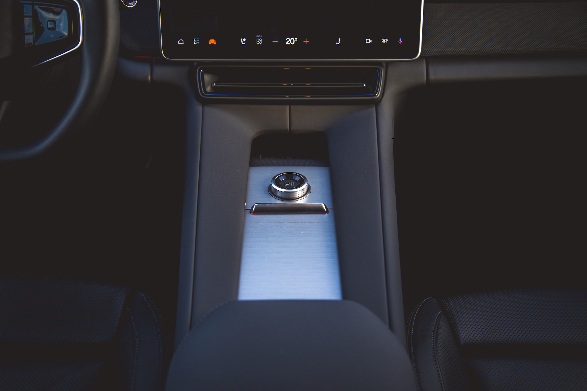 Vista cercana de la consola central de un automóvil moderno que presenta un diseño elegante con una superficie metálica y un dial de control, junto con una pantalla digital que muestra la temperatura y varias configuraciones.