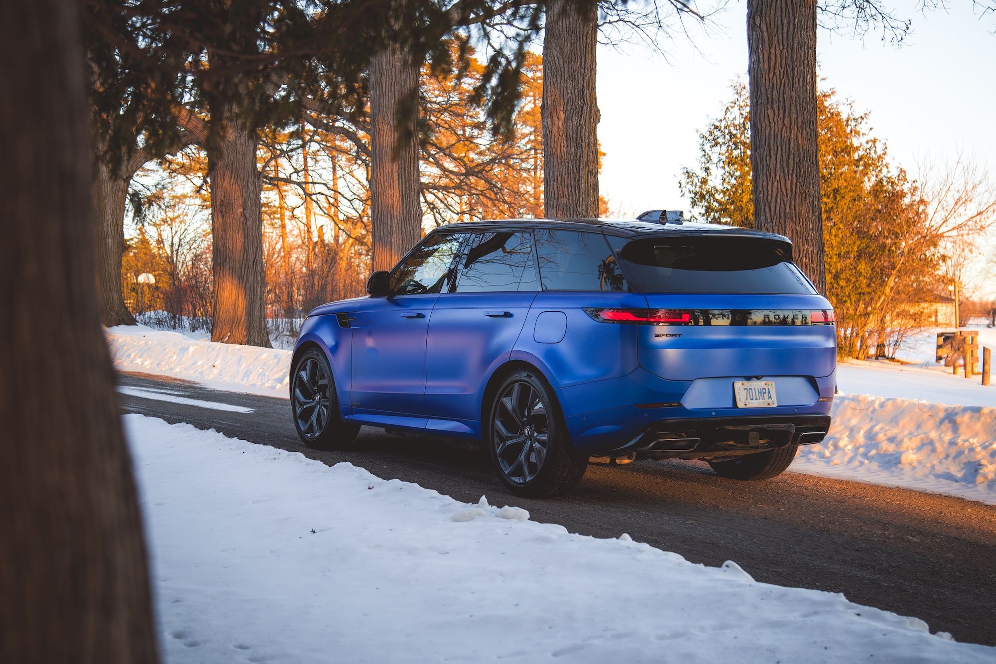 A blue SUV parked on a snow-covered road surrounded by trees during sunset.