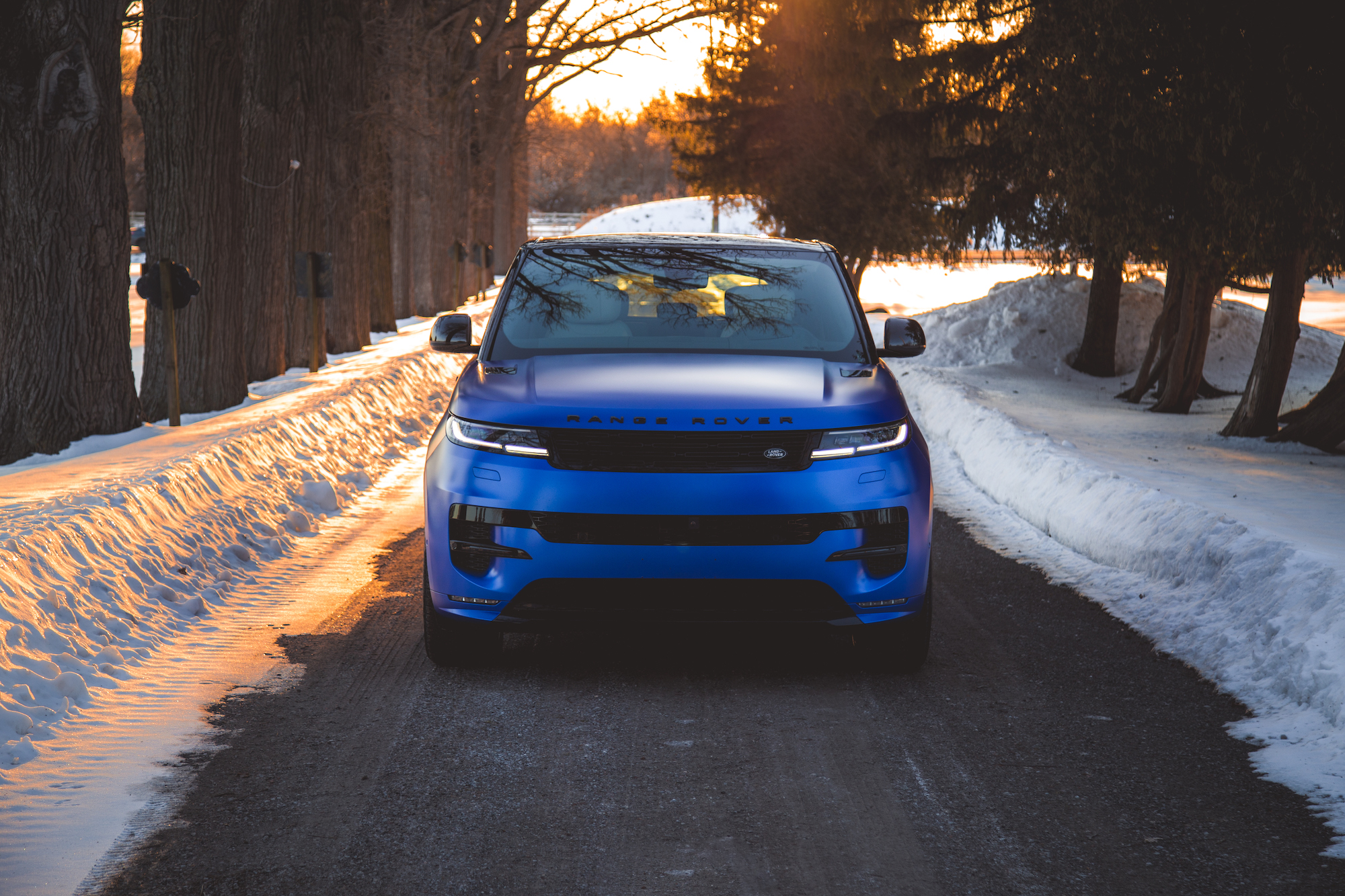 A blue Range Rover parked on a snow-covered road, surrounded by trees at sunset.