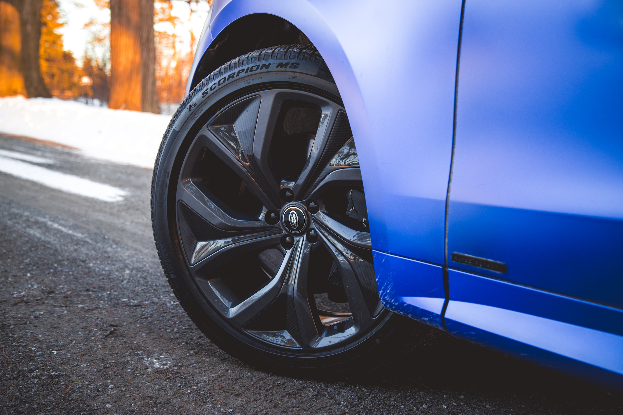 Close-up of a stylish car wheel with a black alloy rim, mounted on a vehicle with a blue exterior, parked on a gravel road beside snow-covered ground.