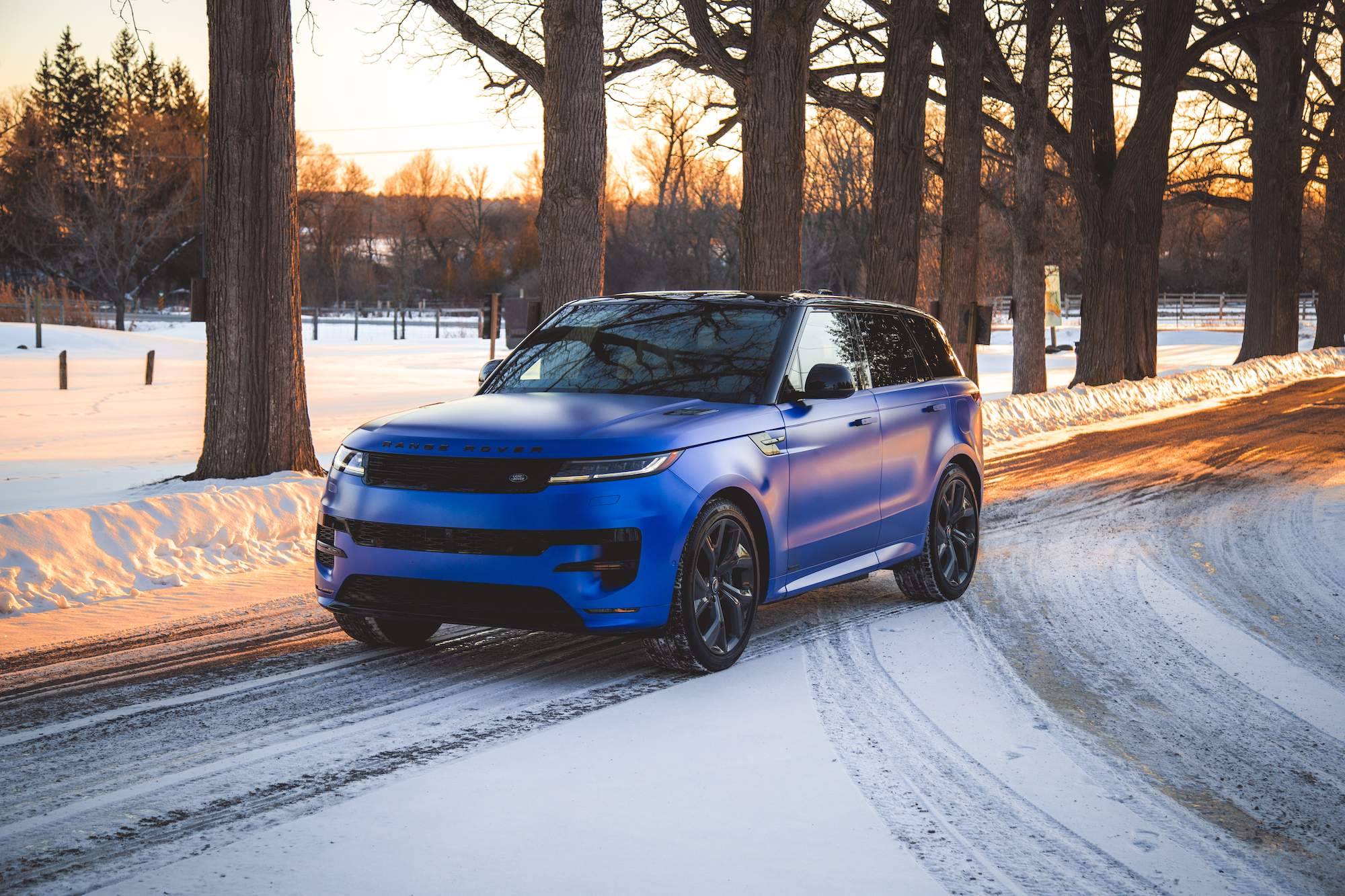 A blue matte Range Rover parked on a snowy road, surrounded by tall trees, with the sun setting in the background.