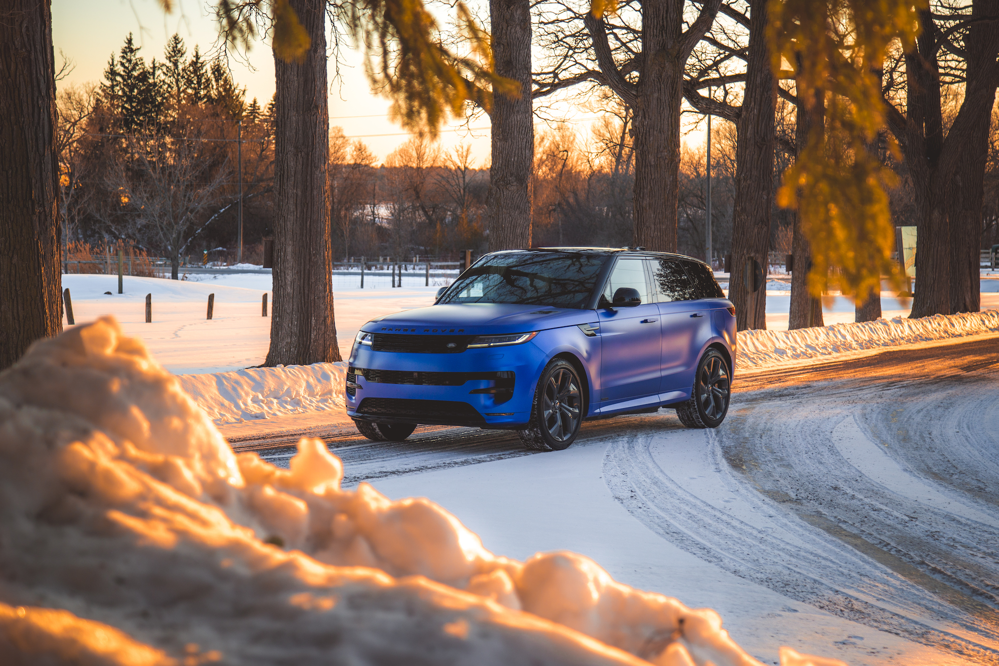 A blue SUV driving on a snowy road surrounded by trees at sunset.