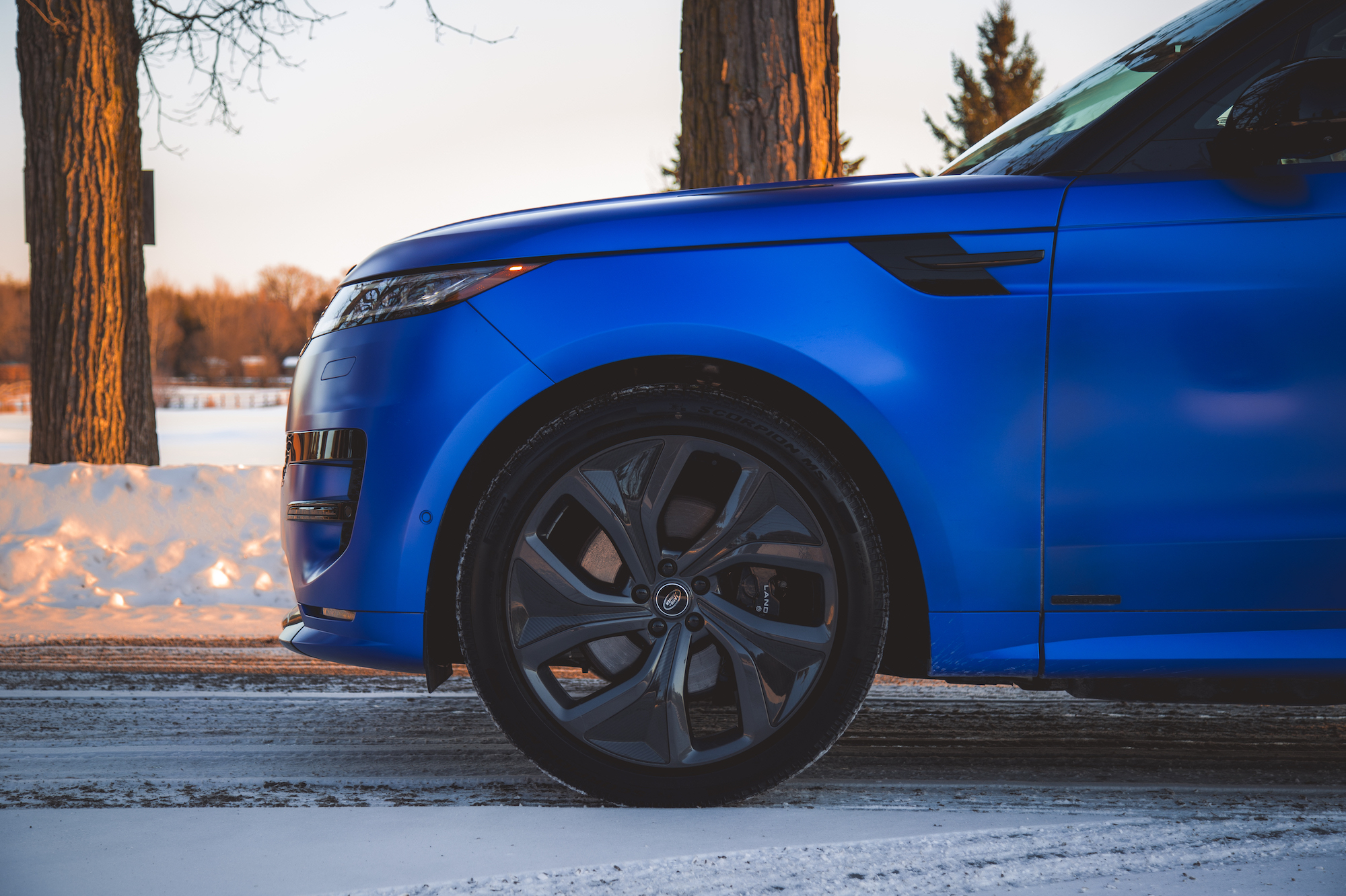 Close-up view of the front side of a blue luxury SUV parked on a snowy road, showcasing its wheel and sleek design.