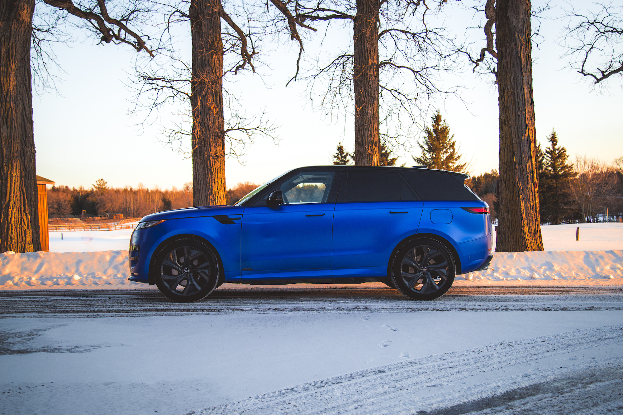 A blue SUV parked on a snowy road, with tall trees and a wooden structure in the background during sunset.
