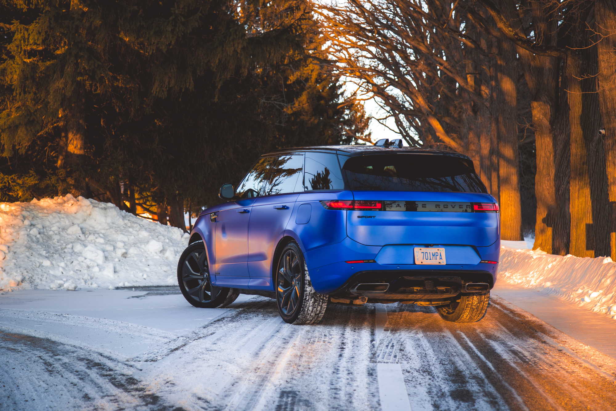 A blue Land Rover Range Rover Sport parked on a snowy road, surrounded by trees at sunset.