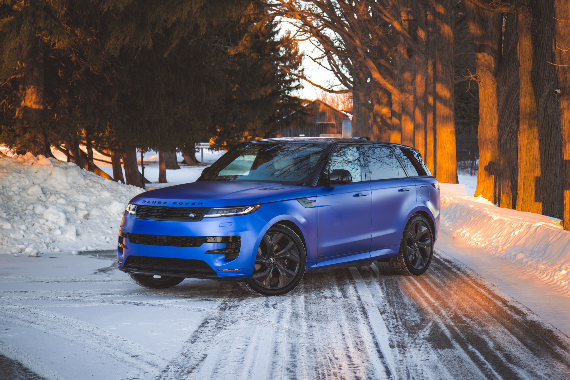 A blue Range Rover parked on a snowy road surrounded by trees, with golden sunlight casting shadows.