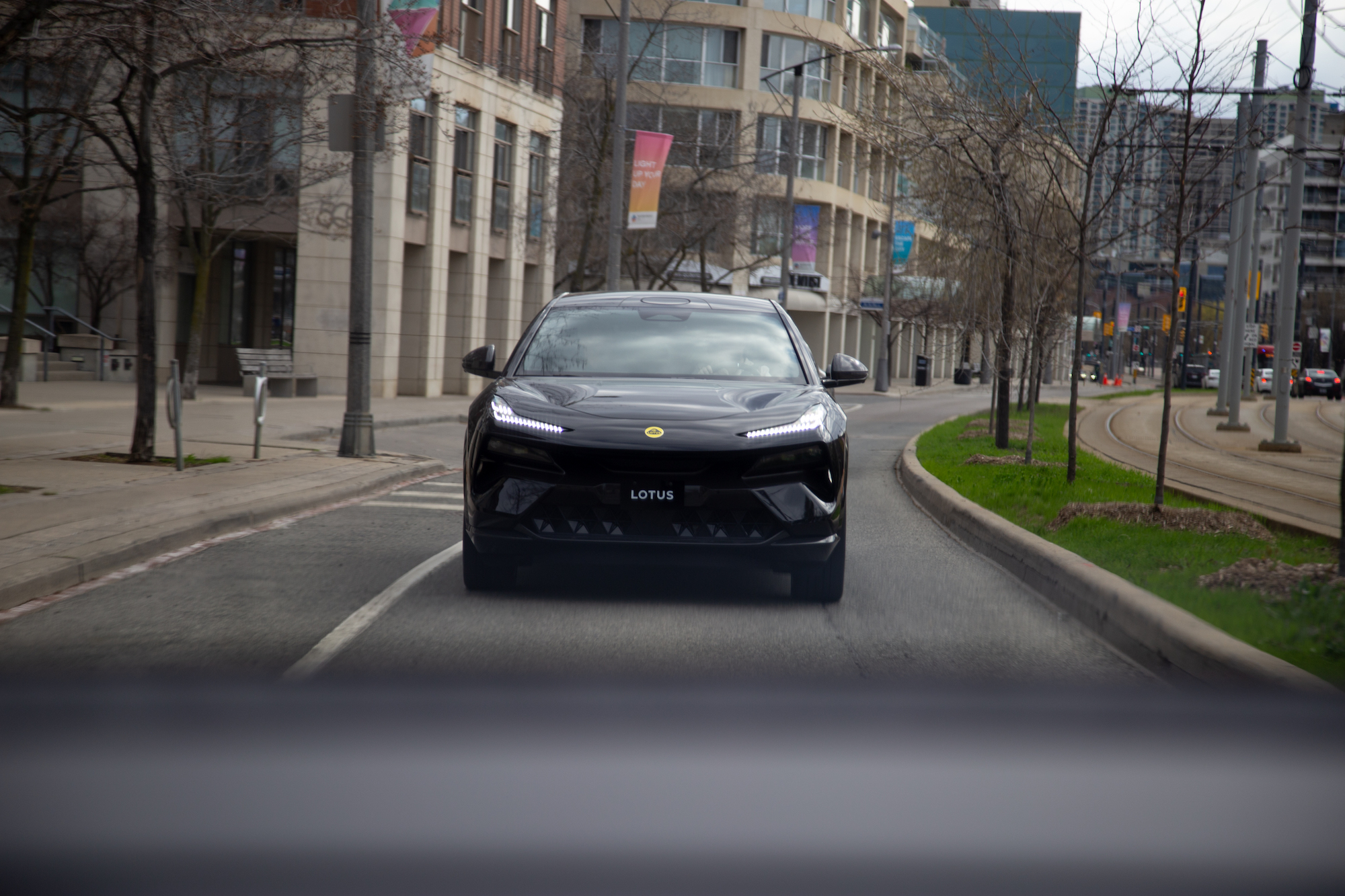 Un automóvil Lotus negro circulando por una calle de la ciudad, con edificios modernos al fondo y árboles sin hojas a lo largo de la carretera.