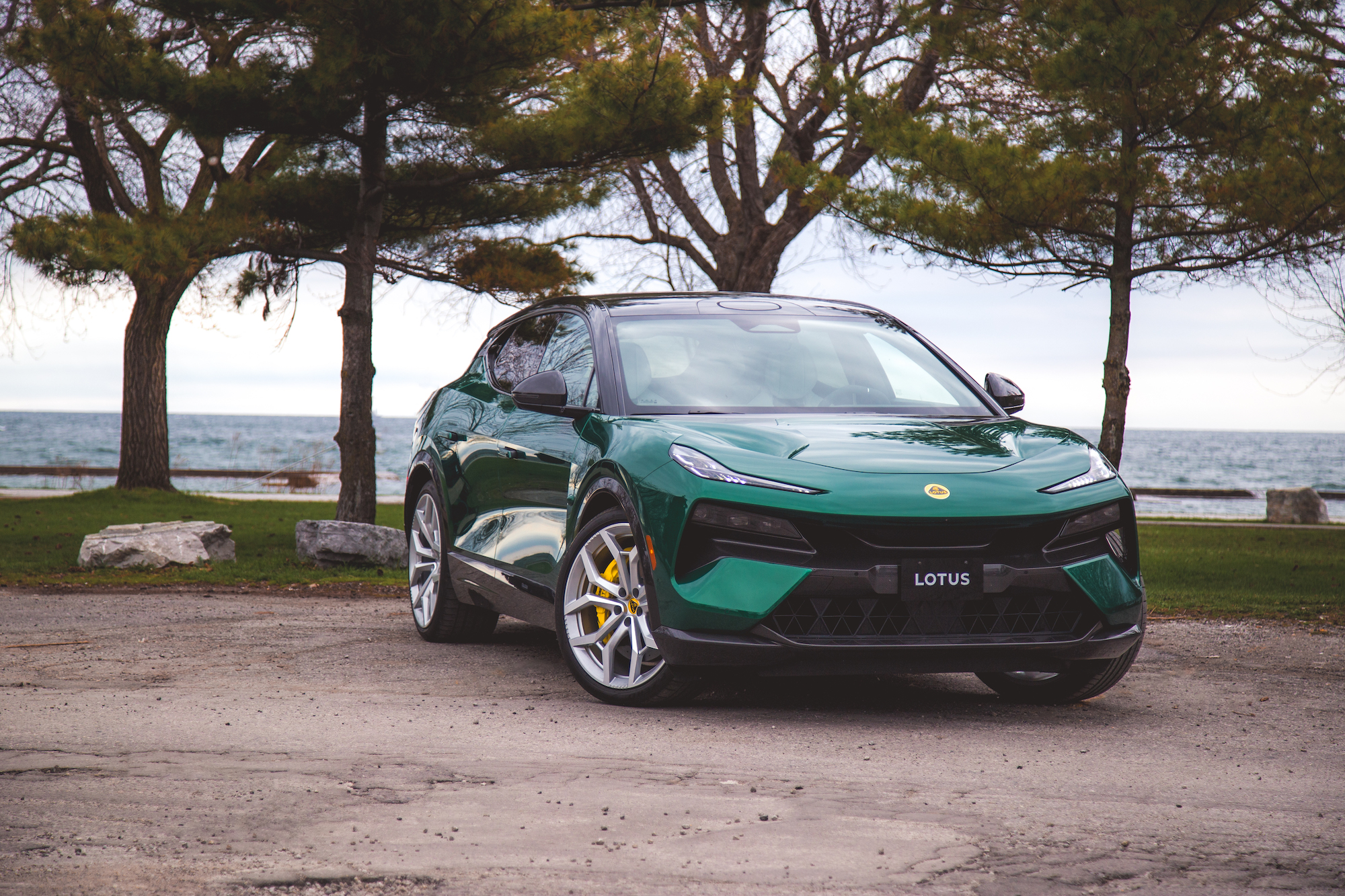 Un SUV Lotus verde estacionado sobre una superficie de grava con un fondo pintoresco de árboles y un lago.