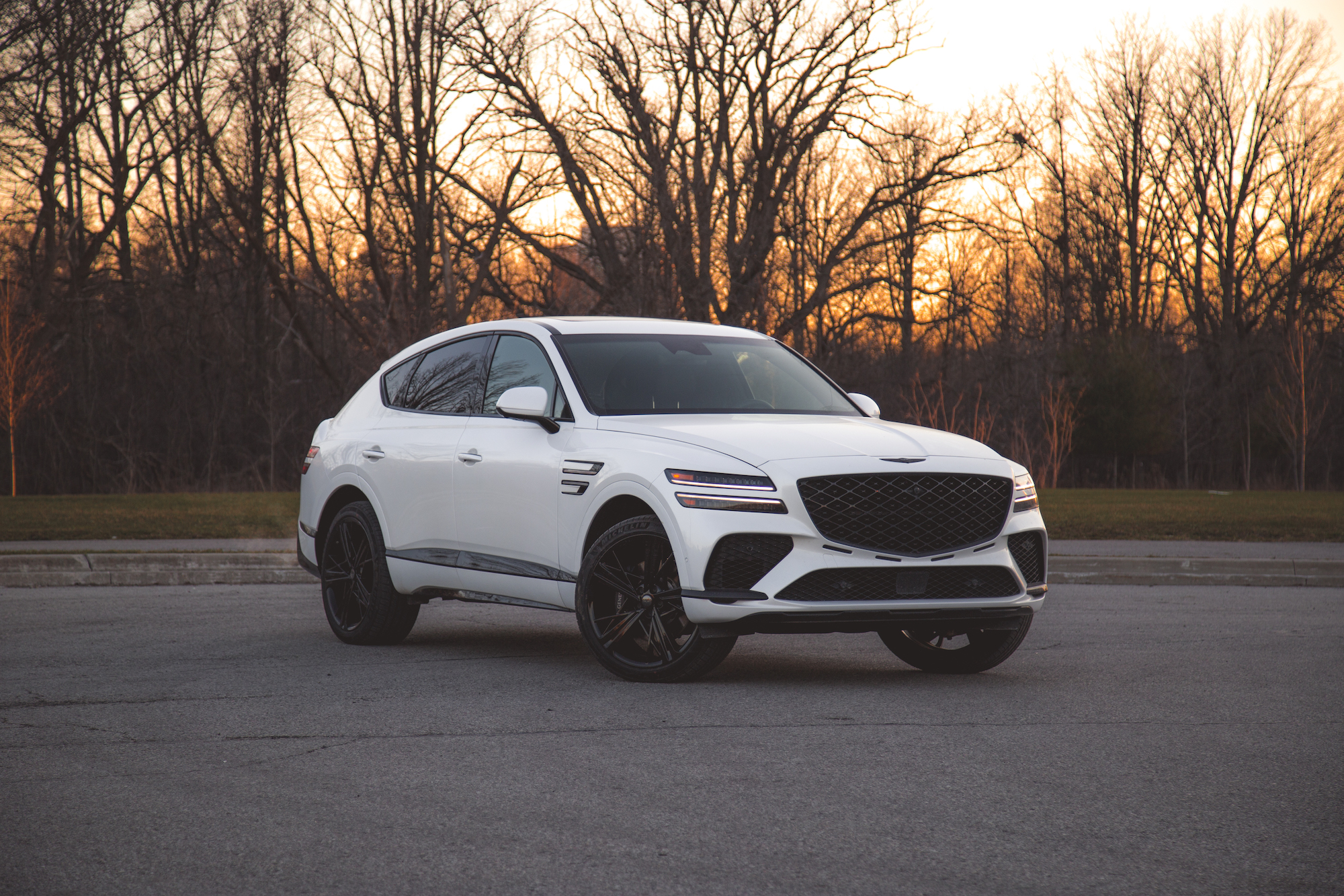 A white luxury SUV parked in an empty lot during sunset, with trees in the background.