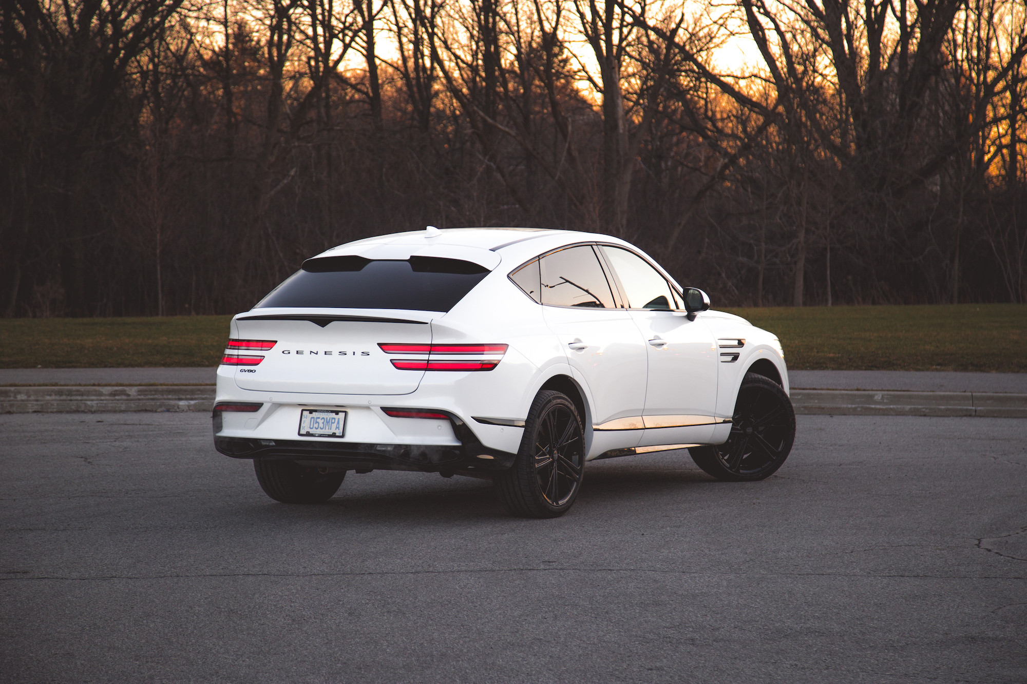 A white Genesis SUV parked on an asphalt surface with a backdrop of bare trees and a sunset sky.