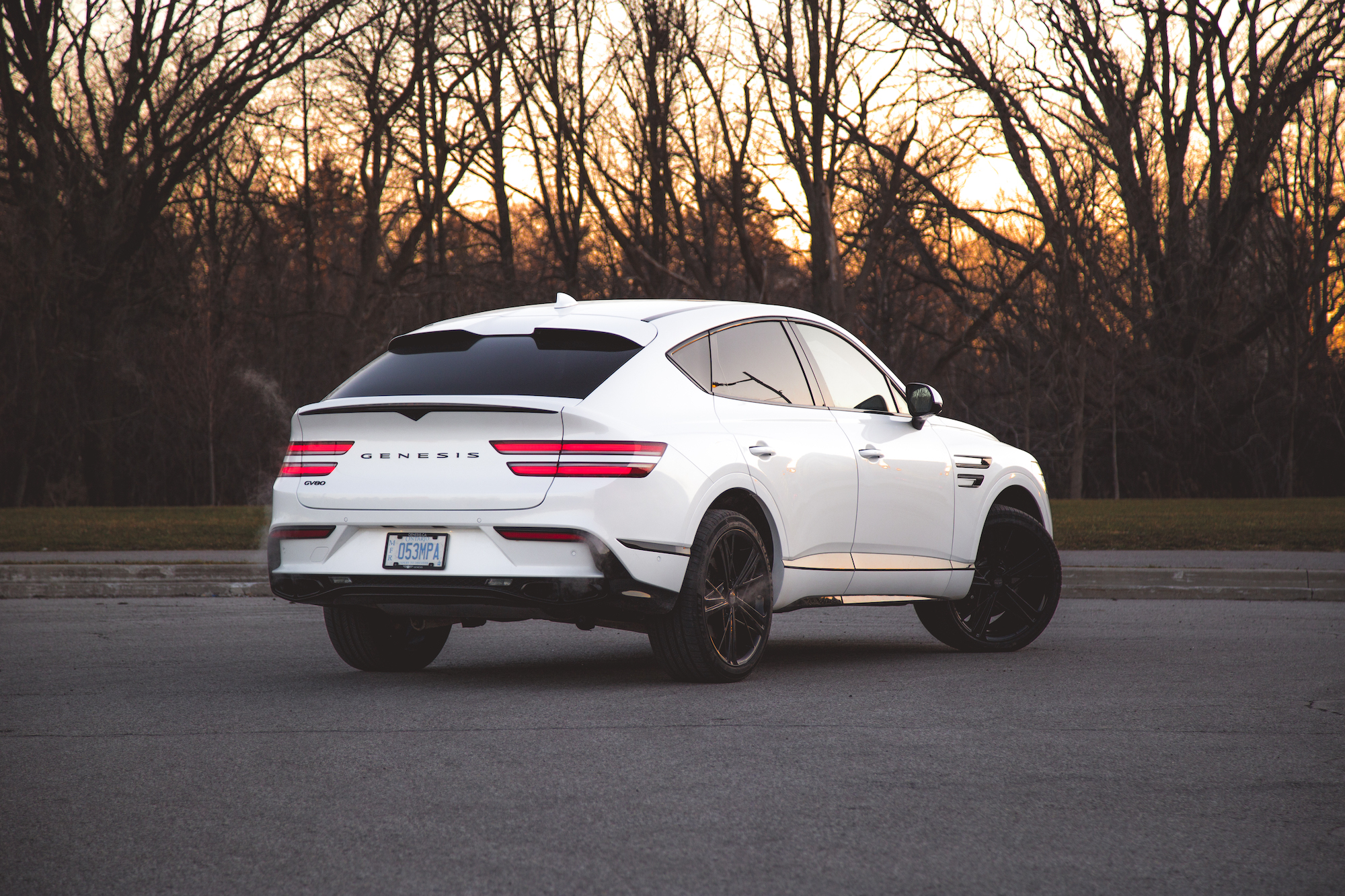 A white Genesis GV70 SUV parked in an empty lot during sunset, showcasing its sleek design and black wheels with a backdrop of bare trees.