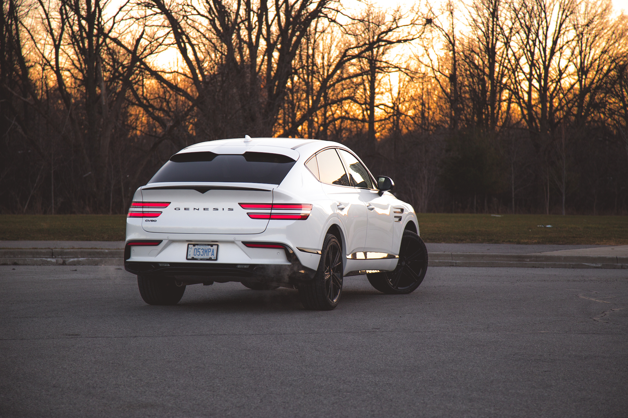 A white Genesis SUV parked on a road at sunset, showcasing its sleek design with black wheels and a clear view of the rear.