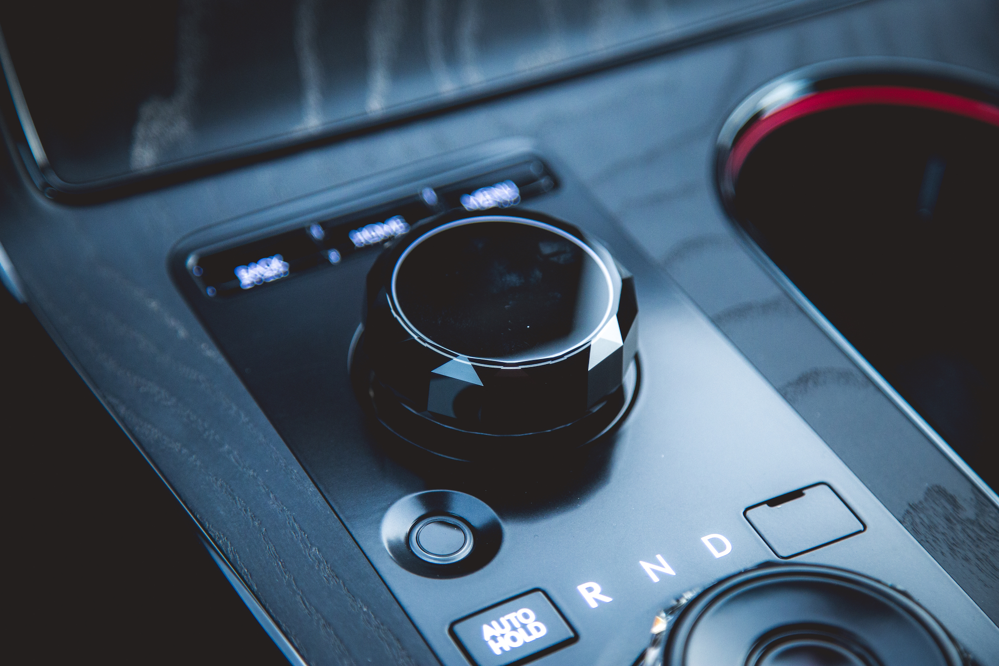 Close-up of a car's gear shift control featuring a black dial, buttons for 'AUTO HOLD' and 'R N D', and a sleek dashboard design.