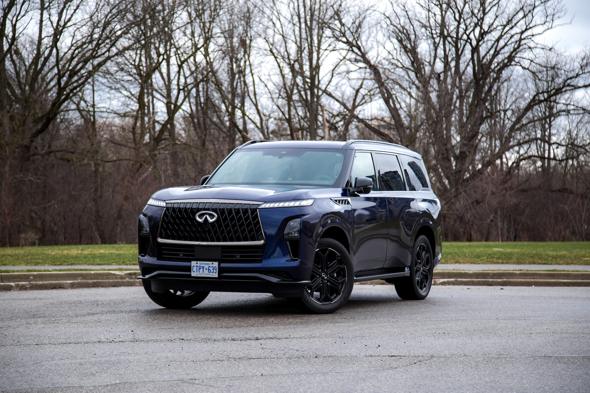 Un SUV de lujo azul oscuro estacionado en una carretera, que muestra un diseño moderno con una parrilla prominente y llantas de aleación negras, con un fondo de árboles desnudos y hierba verde.