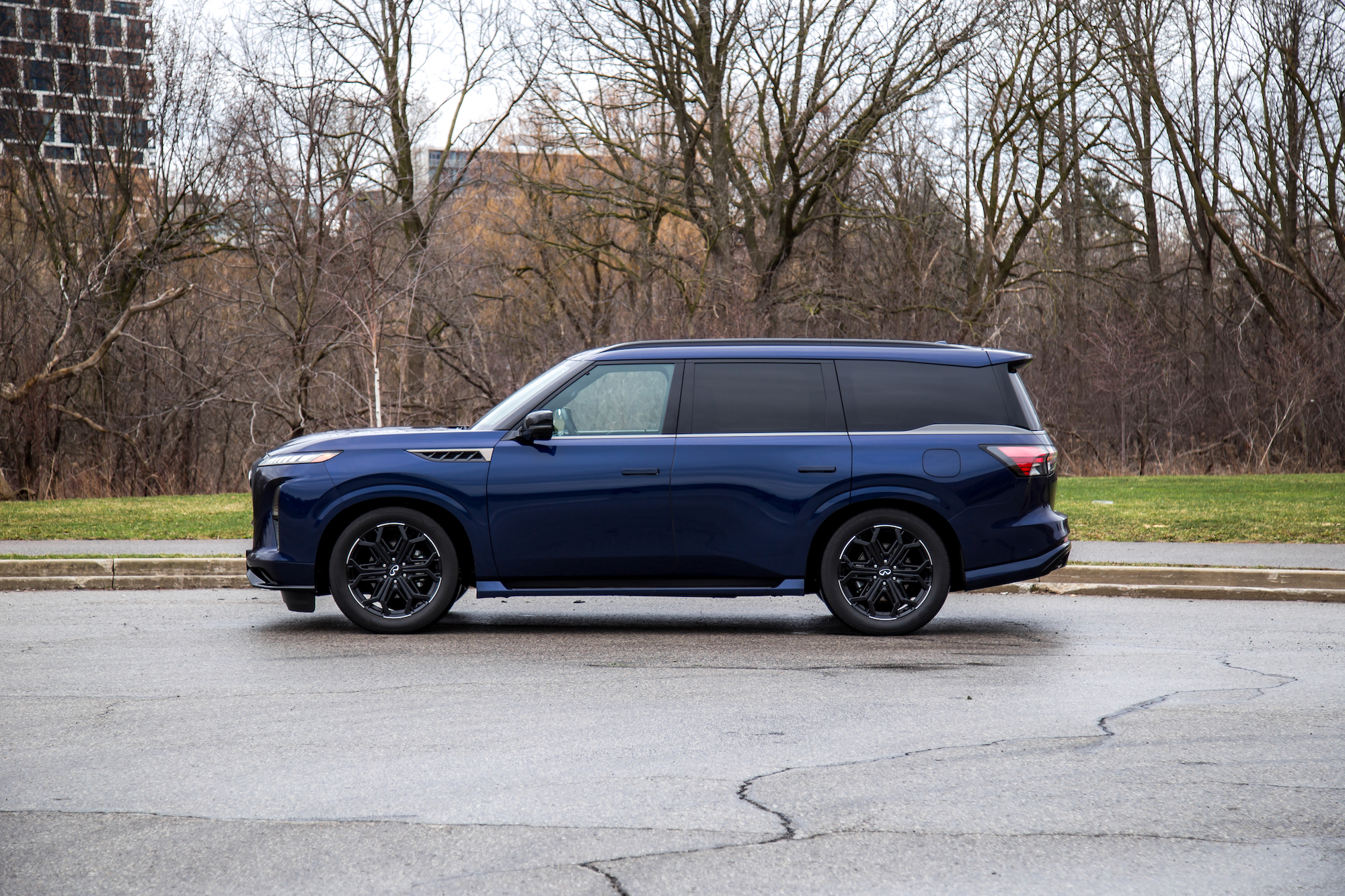 Un SUV azul estacionado en una calle, mostrando su perfil lateral, con un fondo de árboles desnudos y una zona de césped.