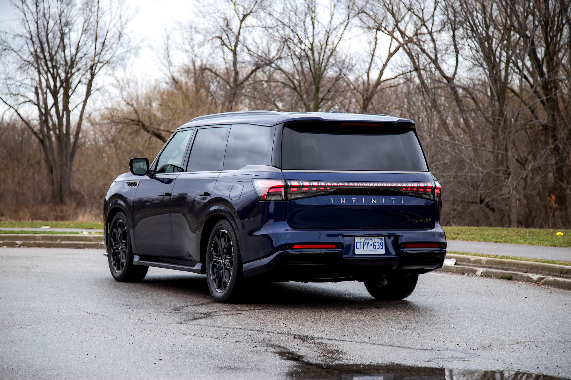 Un SUV Infiniti QX80 azul oscuro estacionado en una carretera mojada con una zona boscosa al fondo.