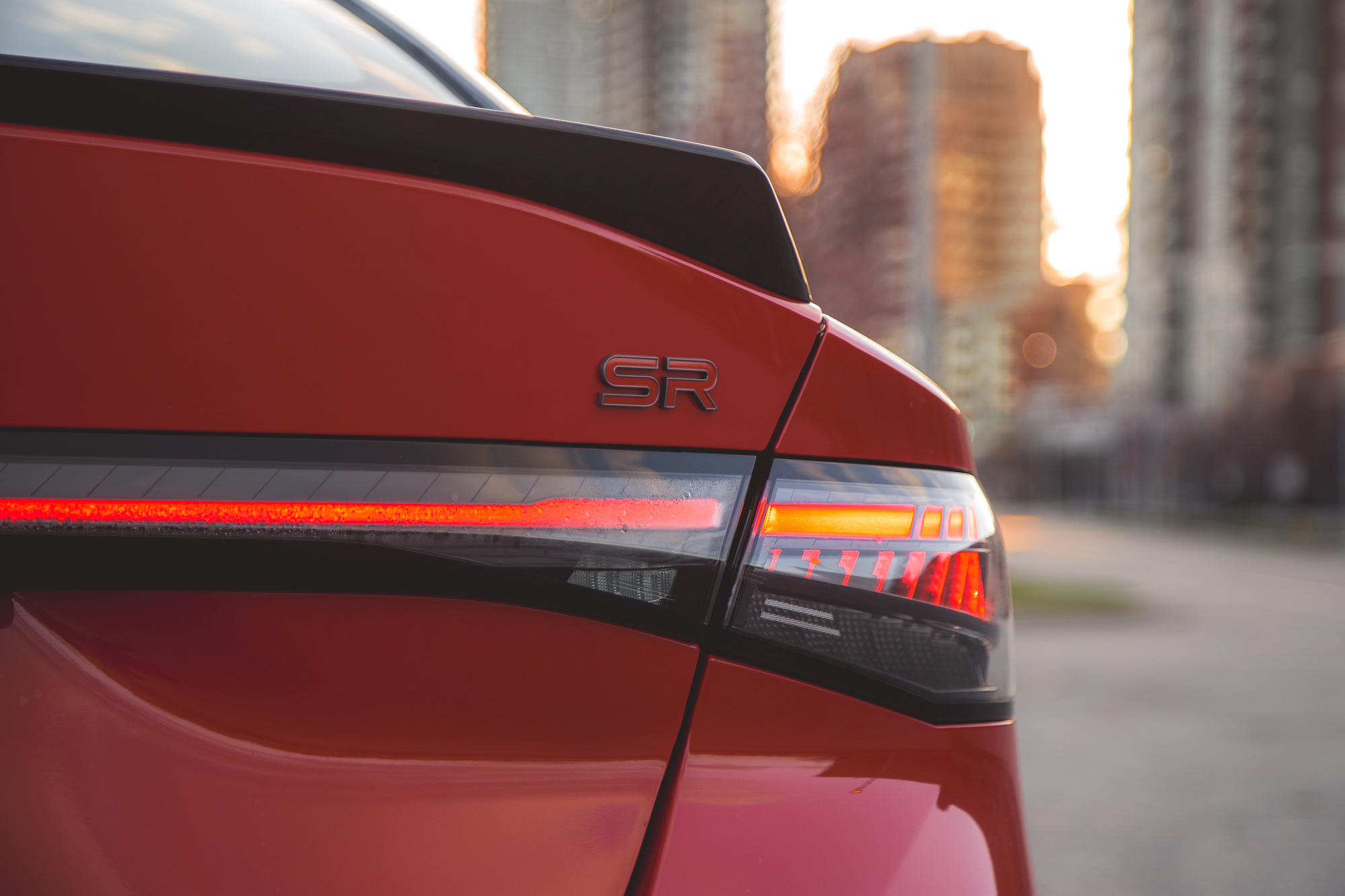 Primer plano de la parte trasera de un automóvil rojo que muestra la insignia 'SR' y las luces traseras, con edificios urbanos al fondo.